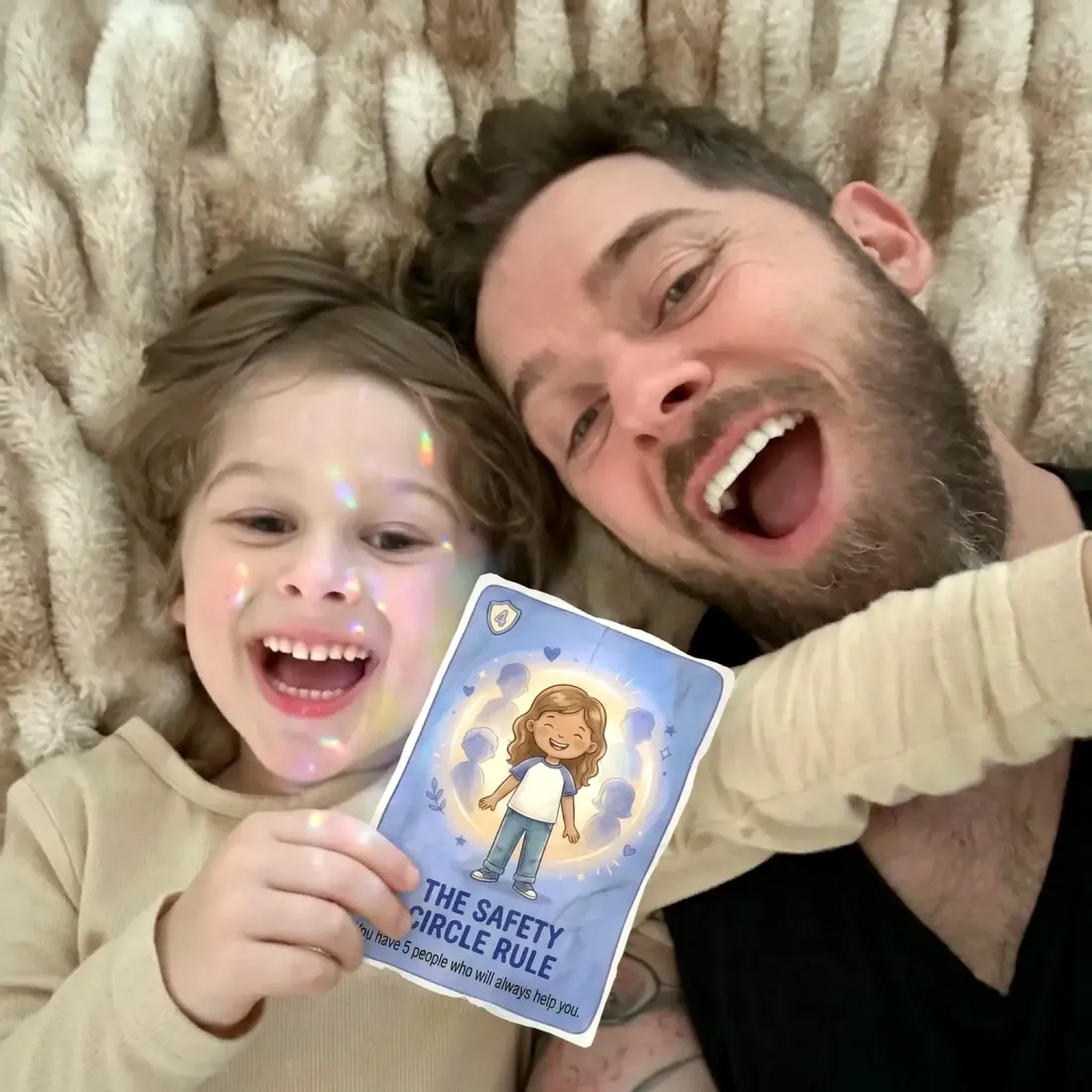A dad and his giggling kid lying on a blanket, holding up the Safety Circle Rule card together